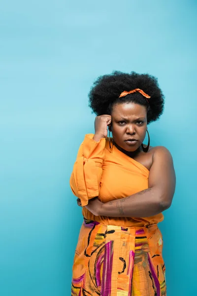Sad african american body positive woman in orange dress and hoop earrings isolated on blue — Fotografia de Stock