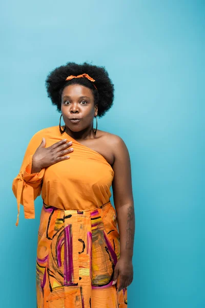 Surprised african american body positive woman in orange dress and hoop earrings isolated on blue - foto de stock
