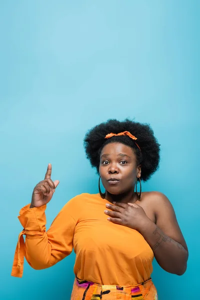 Tattooed african american body positive woman in orange dress and hoop earrings pointing with finger isolated on blue — Stock Photo