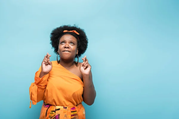 Hopeful african american body positive woman in orange dress and hoop earrings with crossed fingers isolated on blue — Stock Photo