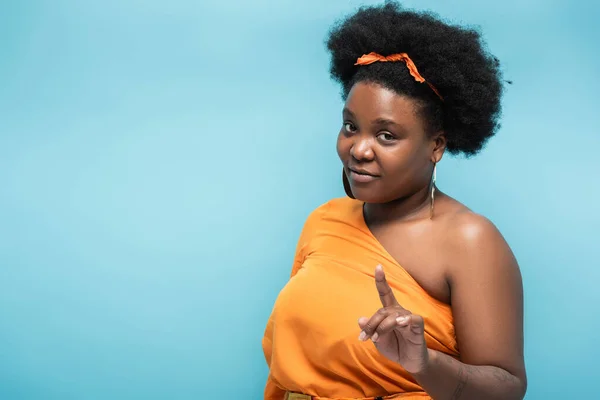 African american body positive woman in orange dress and hoop earrings pointing with finger at camera isolated on blue — Stock Photo