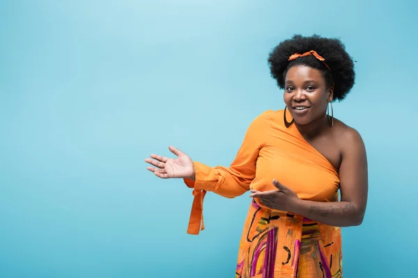 Smiling african american body positive woman in orange dress and hoop earrings pointing with hands isolated on blue — Stock Photo