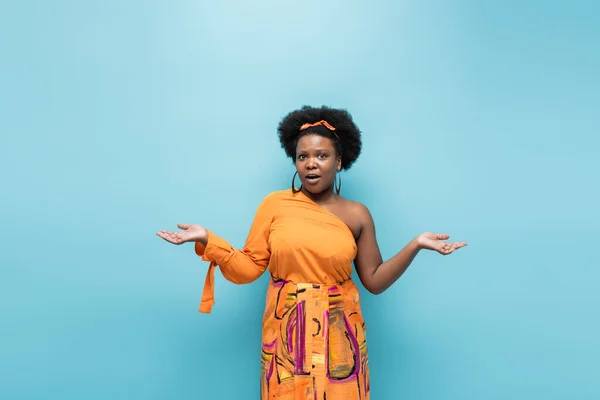 Confused african american body positive woman in hoop earrings showing shrug gesture isolated on blue — Stock Photo