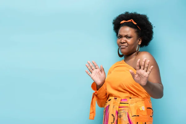 Displeased african american body positive woman in hoop earrings and orange dress showing no gesture isolated on blue — Fotografia de Stock