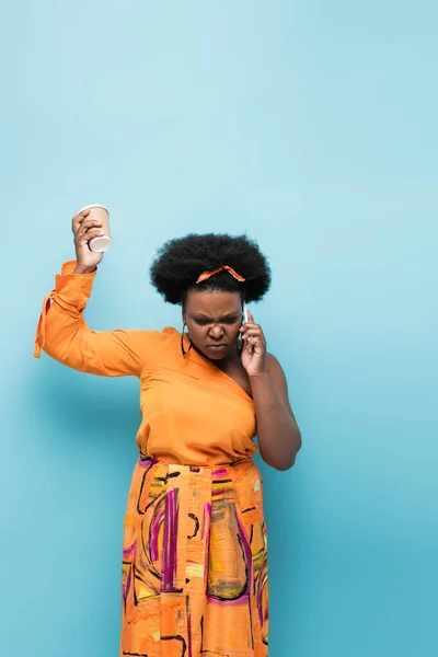 Displeased african american body positive woman holding paper cup and talking on smartphone on blue — Stock Photo