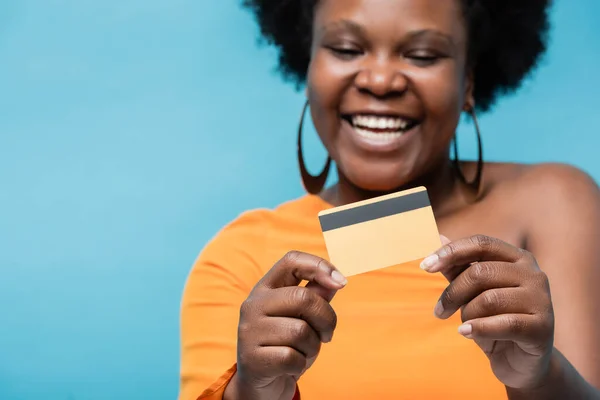 Happy african american body positive woman in hoop earrings holding credit card isolated on blue — Stock Photo