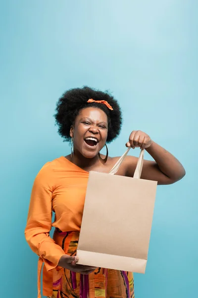 Amazed african american body positive woman in orange dress holding shopping bag isolated on blue — Fotografia de Stock