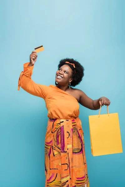 Cheerful african american body positive woman in orange dress holding shopping bag and credit card isolated on blue — Fotografia de Stock