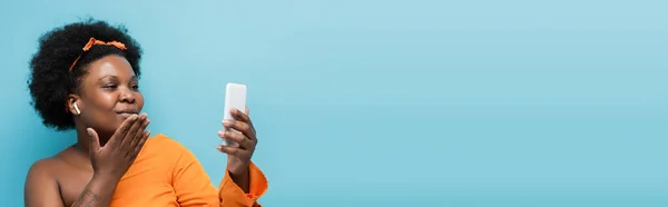 African american body positive woman in wireless earphones having video call and sending air kiss isolated on blue, banner — Stock Photo