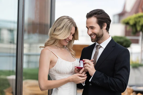 Cheerful man in suit holding box with engagement ring near girlfriend on terrace — Stock Photo