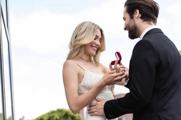 Low angle view of smiling man holding engagement ring near girlfriend in dress on terrace - foto de stock