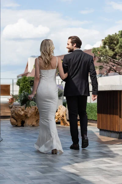 Smiling groom looking at bride in dress with bouquet on terrace of restaurant — Stockfoto