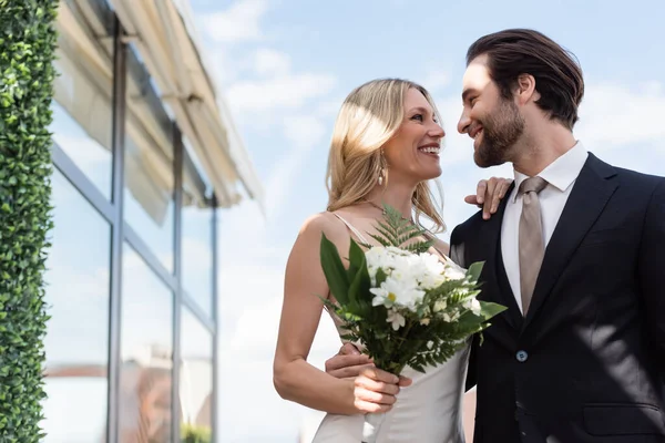 Низький кут зору усміхненої нареченої з квітами, що обіймають нареченого на терасі — стокове фото