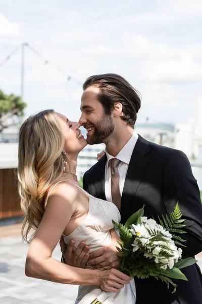 Side view of smiling groom kissing and hugging bride with flowers on terrace — Foto stock