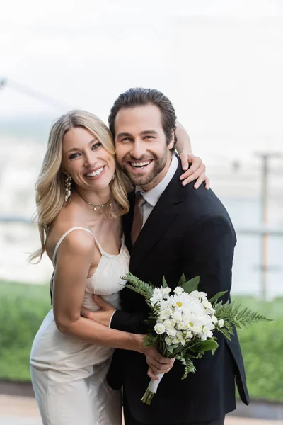 Positive bride in wedding dress holding bouquet and embracing groom on terrace — Stock Photo