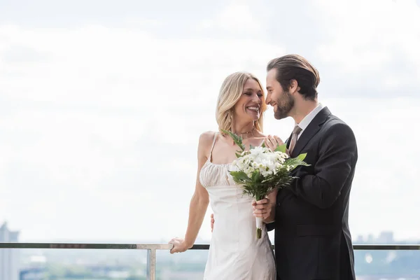 Smiling groom in elegant suit hugging blonde bride with bouquet on terrace — Photo de stock