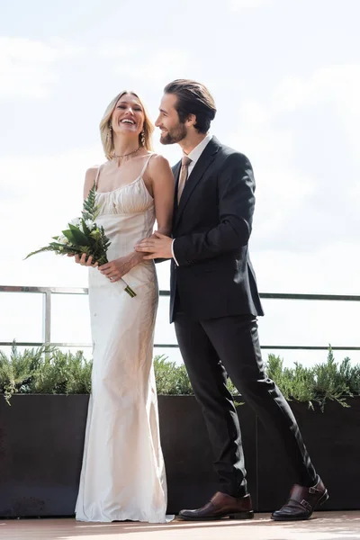 Позитивний наречений в костюмі обіймає наречену в сукні тримає букет на терасі ресторану — стокове фото