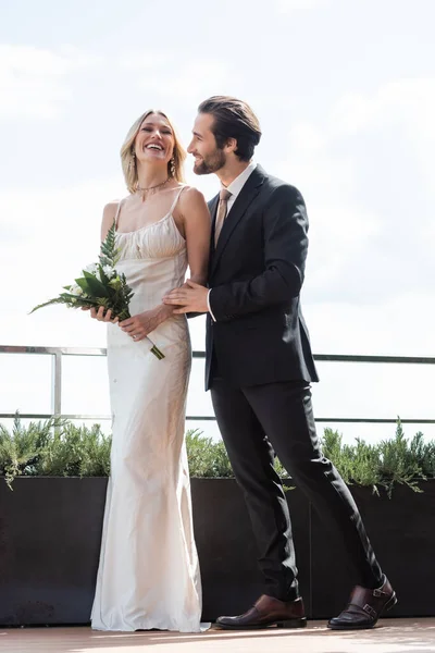 Positive groom in suit hugging bride in dress holding bouquet on terrace of restaurant — Fotografia de Stock