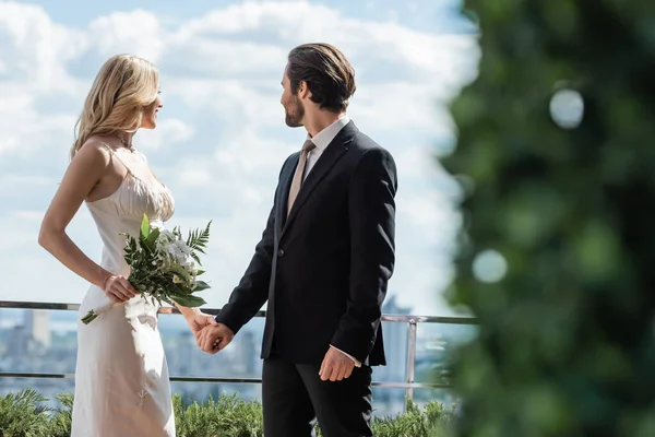 Side view of smiling woman in wedding dress holding bouquet and hand of groom on terrace - foto de stock