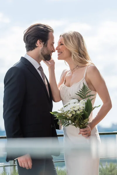 Side view of smiling bride with bouquet touching face of bearded groom on terrace — Stock Photo