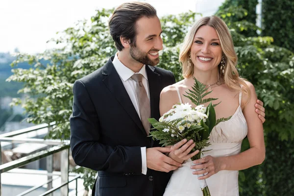 Happy groom in formal wear hugging bride with bouquet on terrace of restaurant — Photo de stock