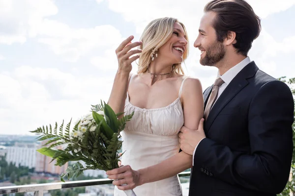 Cheerful bride holding flowers and looking at elegant groom on terrace of restaurant — Fotografia de Stock