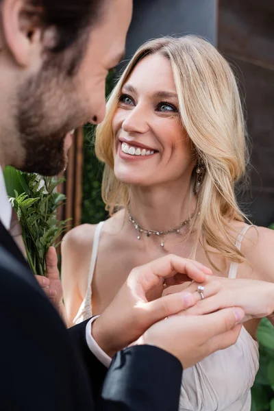 Blurred groom wearing ring on finger of smiling blonde bride outdoors — Stockfoto