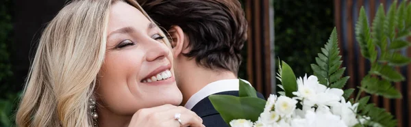 Cheerful blonde bride with flowers hugging groom outdoors, banner — Foto stock