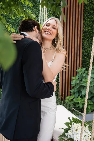 Groom in suit kissing neck of bride near bouquet on terrace of restaurant — Foto stock