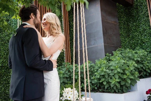Smiling newlyweds embracing near bouquet on swing on terrace — Stockfoto