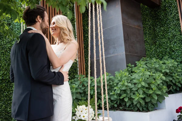 Smiling newlyweds embracing near bouquet on swing on terrace — Photo de stock