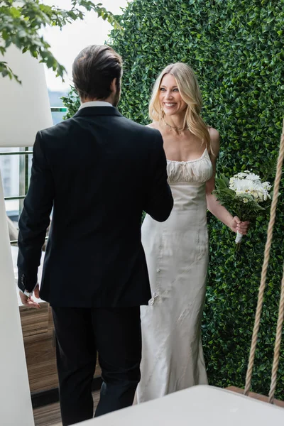 Smiling blonde bride holding bouquet and looking at groom on terrace — Stock Photo