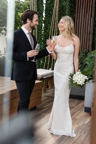 Smiling newlyweds with bouquet and champagne looking at each other on terrace — Stock Photo