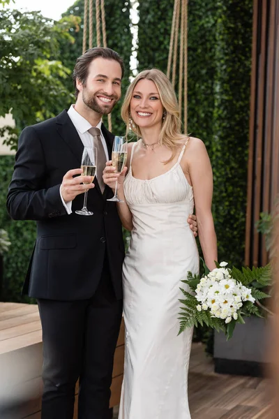 Positive newlyweds with champagne and bouquet looking at camera on terrace — Stockfoto