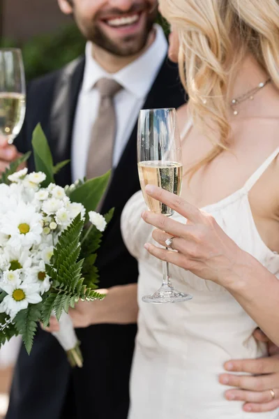 Cropped view of blonde bride holding bouquet and champagne near groom outdoors — Stockfoto