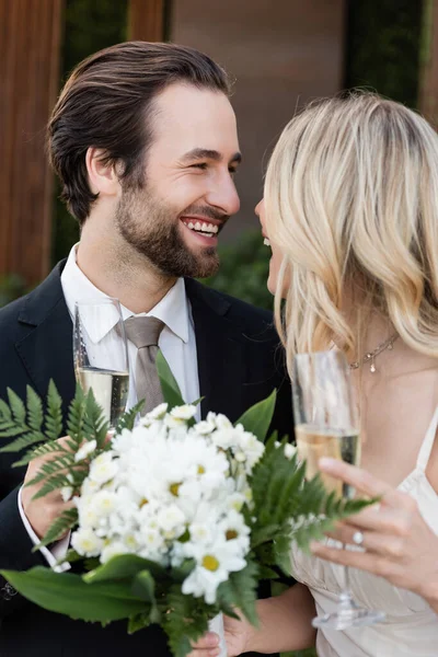 Positive groom holding glass of champagne near bride with bouquet outdoors — Photo de stock