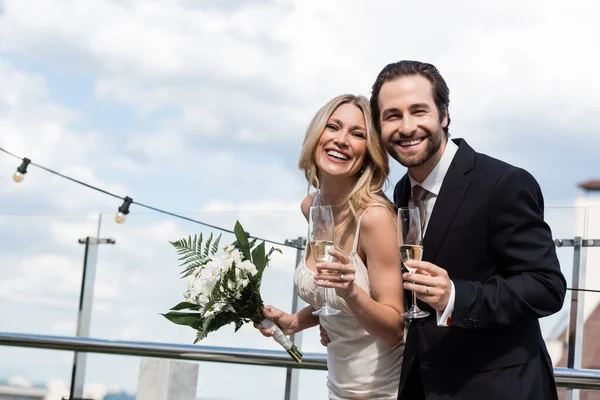 Cheerful newlyweds with bouquet and champagne looking at camera on terrace — Stockfoto