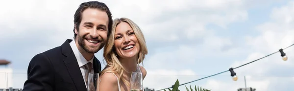 Positive newlyweds with glasses of champagne looking at camera on terrace, banner — Stock Photo