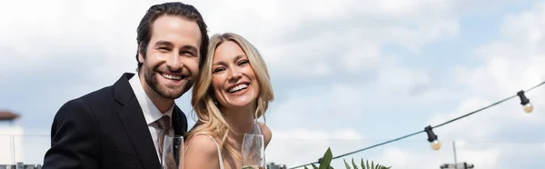 Positive newlyweds with glasses of champagne looking at camera on terrace, banner — Photo de stock