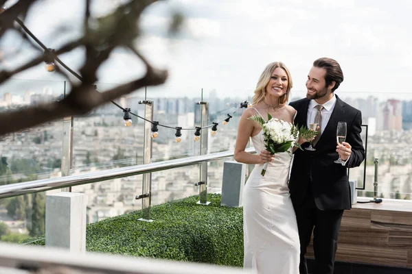 Happy groom holding champagne and looking at blonde bride with bouquet on terrace of restaurant — Photo de stock
