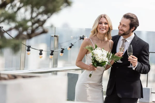 Smiling bride with bouquet and champagne looking at camera near elegant groom on terrace — Stock Photo