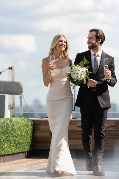 Positive newlyweds with flowers and champagne walking on terrace of restaurant — Foto stock