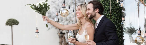 Smiling newlyweds holding champagne and bridal bouquet near light bulbs on terrace, banner — Foto stock