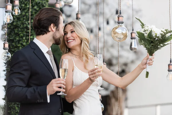 Blonde bride holding bouquet and champagne near elegant groom and light bulbs on terrace — Stockfoto