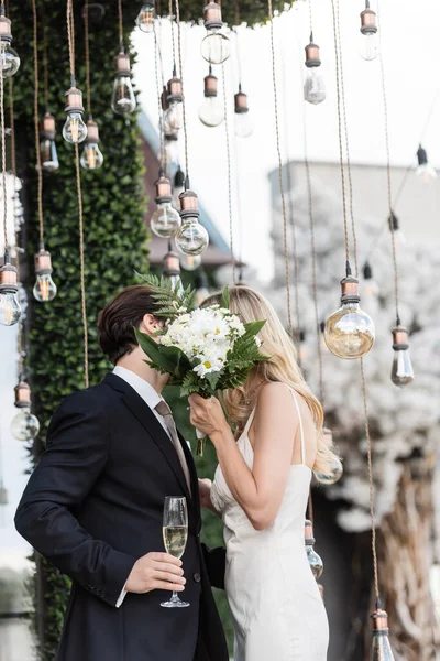 Newlyweds with bouquet and champagne covering faces while kissing under light bulbs outdoors — Foto stock