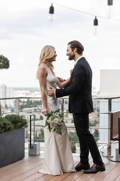 Side view of groom in suit hugging blonde bride with bouquet on terrace — Stock Photo