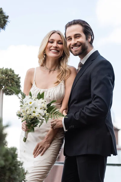 Positive newlyweds with bouquet looking at camera on terrace — Foto stock