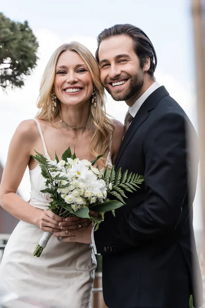 Felice sposi con bouquet sorridente alla fotocamera all'aperto — Foto stock