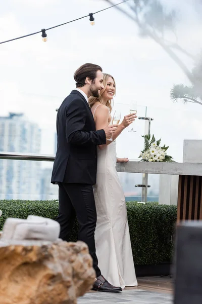 Усміхнений наречений у костюмі тримає келих шампанського біля нареченої та букет на терасі — стокове фото
