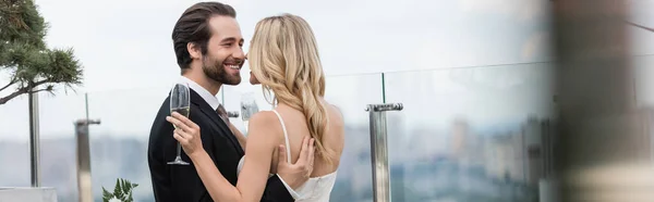 Усміхнений наречений обіймає наречену з шампанським на терасі, банер — стокове фото